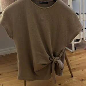 Knyttopp Zara - Säljer en populär beige topp från Zara i mjuk bomullskänsla. Toppen har korta ärmar och en snygg knytning i sidan som ger en cool detalj. Perfekt till jeans eller kjol för en avslappnad men trendig look.