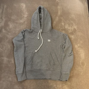 Grå hoodie från Acne Studios - Säljer en stilren grå hoodie från Acne Studios med vit snörning i huvan och diskret logopatch på bröstet. Tröjan har en klassisk känguruficka och mjukt material som gör den skön att bära. Perfekt för chill dagar och enkel att matcha.