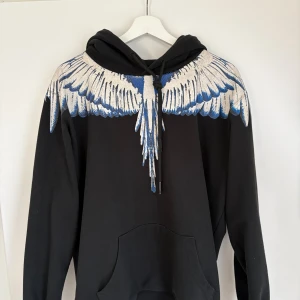 Marcelo Burlon hoodie - Svart hoodie från Marcelo Burlon County of Milan med ikoniskt vingtryck i vitt och blått över axlar och bröst. Tröjan har huva med snörning och stor känguruficka framtill. Perfekt statement-plagg för dig som vill sticka ut.