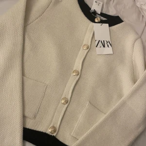 Vit kofta med svarta detaljer från Zara - Stilren vit kofta från Zara med svarta detaljer vid hals och nederkant. Koftan har guldiga knappar framtill, två fickor och är stickad i mjukt material. Perfekt för lager-på-lager och snygg till både jeans och kjol.