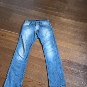 Blå jeans från Nudie Jeans W31 L32 - Säljer ett par klassiska blå jeans från Nudie Jeans med snyggt slitna detaljer och kontrastsömmar. Modellen har raka ben, fem fickor och Nudie Jeans signaturbroderi på bakfickorna. Perfekt passform och cool faded look. 