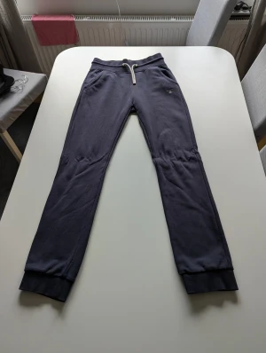 Mörkblå mjukisbyxor från GANT - Snygga mörkblå mjukisbyxor från GANT med broderad logga på höften. Byxorna har ribbade muddar vid bensluten, snörning i midjan och två sidofickor. Perfekta för chill eller häng med kompisar. Mjukt bomullsmaterial för extra komfort.