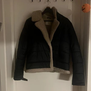 Svart teddyjacka från STR - Svart teddyjacka från STR med beige fuskpäls på krage och insida. Jackan är croppad med bred krage och har quiltade paneler. Perfekt för dig som vill ha en varm och snygg jacka med chill vibe. Stängs med knappar där fram och har två fickor framtill.