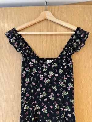 Svart blommig långklänning med volang - Svart långklänning från H&M med små rosa och gröna blommor. Klänningen har volangdetaljer vid axlarna, v-ringad hals och öppen rygg med cut-out. Materialet är lätt och flowy. Slits framtill för extra vibe. Romantisk blommig klänning med öppen rygg, slits, volang axelband och dragkedja i sidan. Storlek XS-S.