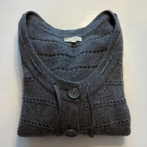 Grå stickad kofta från Jackpot - Mysig grå stickad kofta från Jackpot med snygga dekorativa hål och tre stora knappar framtill. Koftan har ett band i den uringade halsringningen, som går att knyta till rosett, och trekvartsärmar. Perfekt för dig som gillar chill och stilren look.
