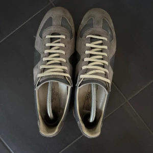 Maison Margiela skor - Maison Margiela sneakers i grå mocka och skinn med beige detaljer och snörning. Snygg retrostil med gummisula och rund tå. Lite slitet bak i sulan. Perfekt för dig som gillar minimalistisk design och vill ha något som sticker ut lite extra. Pris kan diskuteras vid snabbaffär! Tveka inte att höra av dig vid fundering!