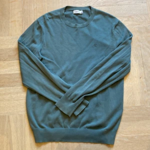 Grön stickad tröja från Calvin Klein - Snygg grön stickad tröja från Calvin Klein med rund halsringning och diskret CK-brodyr på bröstet. Tröjan har långa ärmar och ribbade muddar vid ärmslut och nederkant. Perfekt för lager-på-lager under kyliga dagar.