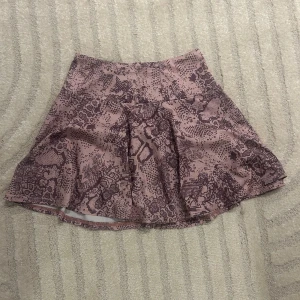 Rosa kortkjol med ormmönster från Coasum - Supersnygg rosa kortkjol från Coasum med coolt ormmönster i mörkare lila toner. Kjolen har en lätt utsvängd form och är gjord i stretchigt material av polyester och spandex. Perfekt för dig som vill sticka ut med ett unikt mönster.