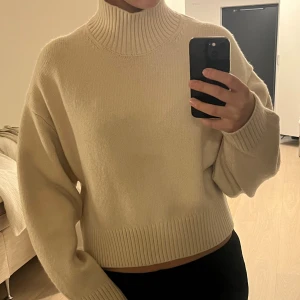 Beige polotröja med ribbad krage - Säljer en beige polotröja med hög, ribbad krage och breda muddar. Tröjan har en loose passform och är stickad i mjukt material, perfekt för kyliga dagar. Enkel och stilren design som är lätt att matcha med olika outfits.