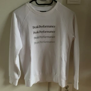 Vit sweatshirt från Peak Performance - Vit sweatshirt från Peak Performance med tryckt logga i olika nyanser på bröstet. Tröjan har rund hals, långa ärmar och ribbade muddar vid ärmslut och nederkant. Perfekt för en clean look.