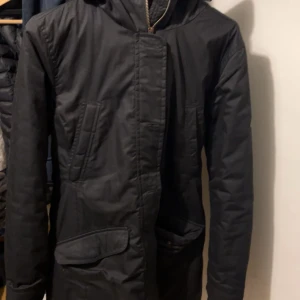 Svart parkajacka från Etirel, strl 40 - Säljer morsans Svart parkajacka från Etirel köpt för ca 1500 men använd 2 ggr max med mjukt, svart foder och quiltad insida. Jackan har dragkedja och knappar framtill, flera fickor och en huva. Materialet är syntet och jackan är perfekt för kalla dagar. Snygg och praktisk design med normal passform.