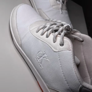 Vita sneakers från Calvin Klein - Passar 40/41/42/43Fräscha vita sneakers från Calvin Klein med snyggt CK-logo broderat på sidan. Skorna har rund tå, snörning och en platt sula med orange detalj undertill. Ovandelen är i slitstark syntet och textil, vilket ger en modern och clean look.