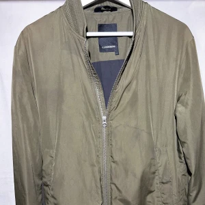 Olivgrön bomberjacka från J.Lindeberg - Snygg olivgrön bomberjacka från J.Lindeberg med klassisk ribbad krage och muddar. Jackan har dragkedja framtill och två sidofickor. Perfekt för dig som gillar stilren och enkel design med en urban touch.