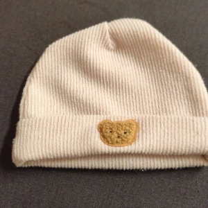 Beige ribbad mössa med nalledekor - Söt beige ribbad mössa med uppvikt kant och en liten broderad nalledekor framtill. Perfekt för dig som gillar gulliga detaljer och vill hålla dig varm med stil. Mjuk och skön passform som funkar till de flesta outfits.