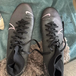 Nike Mercurial svarta fotbollsskor - Säljer ett par Nike Mercurial fotbollsskor i svart med silvriga detaljer och snörning. Skorna har en spetsig tå och platt sula med dobbar för gräsplan. Ovandelen är i syntetmaterial med ett mönstrat, nästan pixeligt utseende och Nike-logga på sidan.