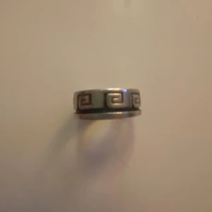 Silver Ring - Size 10 (2cm diameter, ~7.5cm omkrets). Cool bred ring i silverfärg med ett grafiskt mönster som går runt hela ringen. Mönstret är inspirerat av antika grekiska former och ger en unik vibe. Perfekt för dig som gillar statement-smycken och vill sticka ut lite extra.