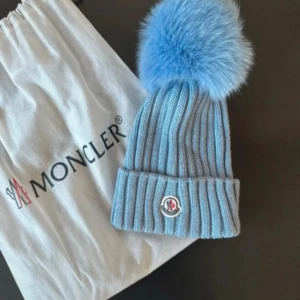 Blå Moncler mössa med pälsboll - Ribbstickad ljusblå mössa från Moncler med stor fluffig pälsboll i matchande blått. Mössan har uppvikt kant och Monclers klassiska logga framtill. Perfekt accessoar för kalla dagar med extra stilpoäng.
