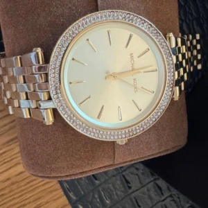Guldig Michael Kors klocka  - Snygg guldig klocka från Michael Kors med rund urtavla och glittrande stenar runt boetten. Länken är i metall med polerad finish och urtavlan har minimalistiska markeringar. Perfekt accessoar för dig som gillar stilrena detaljer och lite extra bling.