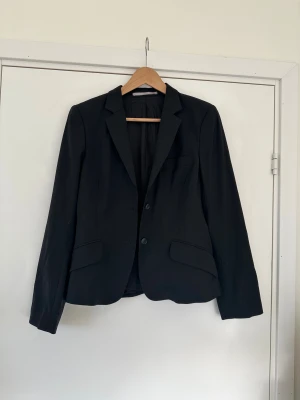 Svart kavaj från Tiger of Sweden - Klassisk svart kavaj från Tiger of Sweden med två knappar framtill, fickor med lock och snyggt figursydd passform. Fodrad insida med diskret mönster och märkets logga. Perfekt för en stilren look. storlek 38. 