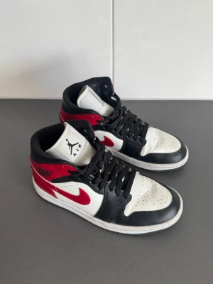 Nike Air Jordan 1 Mid - Nike Air Jordan 1 Mid sneakers med klassisk design i rött, svart och vitt. Skorna har snörning, perforerad tå och rund sula. Swoosh-logga i rött på sidan och Air Jordan Wings på ankeln. Tillverkade i skinn med svart foder och vit platt sula.