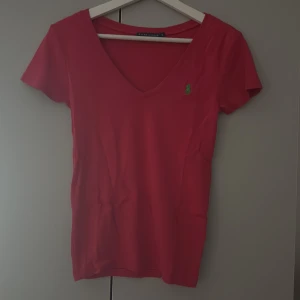Röd v-ringad topp från Ralph Lauren - Säljer en mörk rosa  v-ringad topp från Ralph Lauren med korta ärmar och den klassiska gröna logon broderad på bröstet. Toppen är i mjuk bomull och har en slim passform som sitter snyggt. Perfekt för dig som gillar stilrena och sportiga plagg.