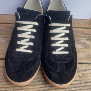 Maison Margiela svarta sneakers  - Maison Margiela sneakers i svart mocka med kontrasterande vita snören och beige gummisula. Klassisk rund tå och platt sula, med märkets ikoniska nummerdetalj på tungan och sula. Snygg retrostil som sticker ut med lyxig känsla. Kvitto finns, nytvättade
