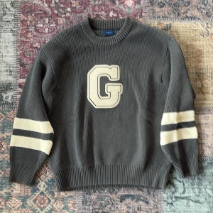 Grå stickad tröja med GANT-logga - Snygg grå stickad tröja från GANT med stor beige G på bröstet och coola vita ränder på ärmarna. Tröjan har rund hals och ribbade muddar vid ärmslut och nederkant. Perfekt för dig som gillar klassisk college-stil.