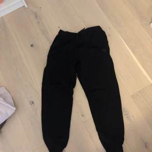 Svarta mjukisbyxor från Craft - Svarta mjukisbyxor från Craft i storlek S. Byxorna har ribbade muddar vid bensluten och snörning i midjan för skön passform. Perfekta för chill eller träning, med diskret Craft-logga på höften. Mjukt material som känns bekvämt hela dagen.