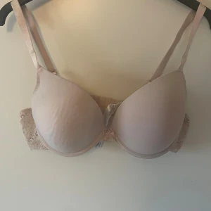 Beige push-up bh från Censored - Säljer en beige push-up bh från Censored i storlek 90D. Bh:n har släta kupor, justerbara axelband och spetsdetaljer på sidorna samt en liten rosett framtill. Perfekt för dig som vill ha en diskret och bekväm bh med snygga detaljer.