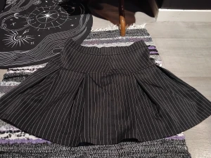 Svart kortkjol med vita ränder - Snygg svart kortkjol med vita tunna ränder i klassiskt pinstripe-mönster. Kjolen har en markerad midja och utställd form med fina veck som ger extra volym. Perfekt för dig som gillar en stilren men edgy look. Står L på den, men är mer M