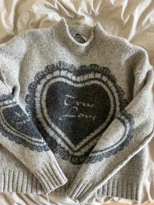 Grå stickad tröja Acne Studios hjärta - Supermysig grå stickad tröja från Acne Studios med stort mörkgrått hjärtmotiv och texten 'True Love' på bröstet och ärmen. Tröjan har hög krage, ribbade muddar och loose fit. Perfekt för dig som gillar coola detaljer och unik design.