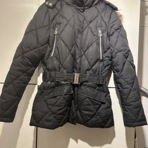 Svart quiltad dunjacka från Röhnisch - Svart dunjacka från Röhnisch med quiltat mönster, huva med fuskpäls och snyggt bälte i midjan. Jackan har dragkedjor på fickorna och detaljer i silver. Perfekt för kalla dagar med sin värmande och vind- samt vattenavvisande design.