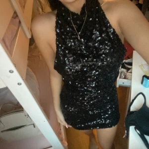 Svart paljettklänning med halterneck - Supersnygg svart kort paljettklänning med halterneck och figurnära passform. Klänningen är täckt av glittrande paljetter som ger en riktigt festlig look. Perfekt för dig som vill sticka ut och glänsa på nästa party!
