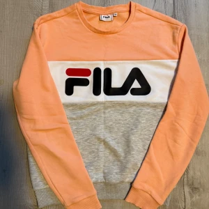 FILA sweatshirt i persika och grå - Säljer en snygg sweatshirt från FILA i persikofärg med grå och vit panel samt stor logga framtill. Tröjan har rund halsringning och långa ärmar. Perfekt för dig som gillar sportig och trendig stil. Passar xs- s storlek. Kan sammfrakta 