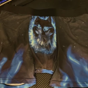 Grisch kallingar - Unika svarta shorts med blå eldliknande detaljer och ett stort vargtryck framtill. Stretchigt material och midwaist passform, perfekt för dig som gillar djurmotiv och coola prints. Snygg kontrast mellan mörka och ljusa färger, riktigt statement-plagg. Sjukt laidback och grisch