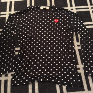 Svart prickig långärmad tröja CdG - Svart långärmad tröja från Comme des Garçons PLAY med vita prickar och det ikoniska röda hjärtat på bröstet. Slimmad passform och mjukt bomullsmaterial som känns skönt mot huden. Perfekt för dig som vill sticka ut med en klassisk men lekfull look.