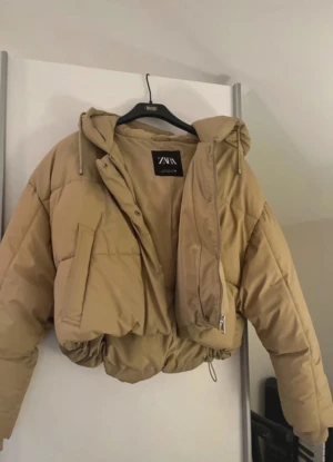 Beige croppad pufferjacka från Zara - Säljer en beige croppad pufferjacka från Zara med huva och snörning i nederkant. Jackan har dragkedja framtill, stora fickor och en loose passform. Perfekt för dig som gillar streetstyle och vill ha en varm jacka med cool look.