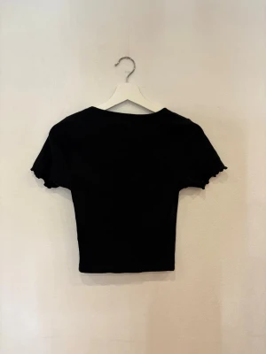 Svart croppad topp från Bik Bok - Svart croppad topp från Bik Bok med korta ärmar och v-ringad hals. Toppen har ribbad struktur och en snygg rynkning framtill vid halsen. Ärmsluten har en vågig kant som ger en extra detalj. Perfekt att styla med jeans eller kjol.