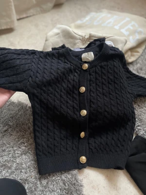 Svart kabelstickad kofta med guldknappar - Svart kabelstickad kofta med rund hals och fem snygga guldknappar framtill. Tröjan har ribbade muddar vid ärmslut och nederkant, vilket ger en schysst passform. Perfekt för dig som gillar klassisk stil med en twist.