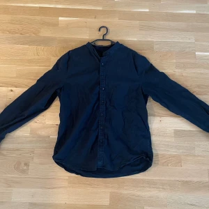 Svart henley-skjorta i bomull - Svart henley-skjorta med lång ärm och knappar hela vägen framtill. Skjortan har en enkel, kraglös design och är tillverkad i mjuk bomull. Perfekt för en avslappnad och stilren look. Har bara tagit bort lappen med storleken. Därav ett litet märke som syns på bilden