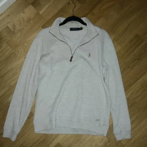 Beige half zip tröja Polo Ralph Lauren - Snygg beige långärmad tröja från Polo Ralph Lauren med klassisk broderad logga på bröstet. Tröjan har en stilren krage och half zip-dragkedja med läderdetalj. Perfekt för dig som gillar enkel och clean stil.