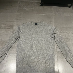 Grå långärmad tröja i bomull - En enkel och stilren grå långärmad tröja med rund hals. Tröjan är tillverkad i mjuk bomull och har ribbade muddar vid ärmslut och nederkant. Perfekt för chill dagar eller när du vill ha en clean look.