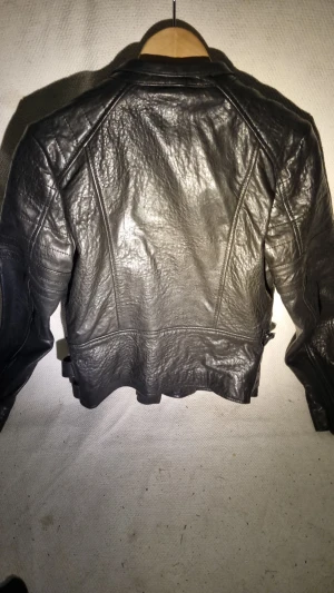 Svart skinnjacka Hollies Stockholm 38 - Cool svart skinnjacka från Hollies Stockholm i äkta fårskinn. Jackan har flera dragkedjor, snygga detaljer med bälte i midjan och klassisk bikerstil. Insidan är fodrad med guldig satin för extra lyxig känsla. Perfekt för dig som gillar edgy och stilrent.