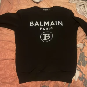 Svart sweatshirt från Balmain Paris - Svart sweatshirt från Balmain Paris med vit logotyptryck på bröstet och rund hals. Tröjan har långa ärmar och en klassisk passform, perfekt för dig som gillar stilrena och trendiga märkesplagg.