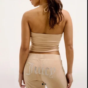 Beige Juicy Couture mjukisbyxor - Säljer ett par ikoniska beige mjukisbyxor från Juicy Couture med strassdetaljer som stavar 'Juicy' över bakfickan. Byxorna har snörning i midjan och är tillverkade i mjuk velour med en loose och wide passform. Perfekta för en chill och trendig look. Nypriset är 1 200 kr!