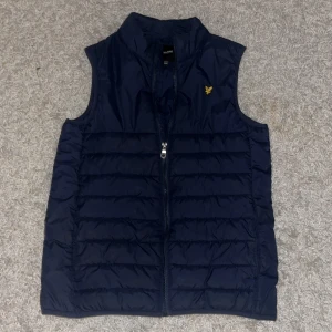 Marinblå dunväst från Lyle & Scott - Snygg marinblå dunväst från Lyle & Scott Junior med gul broderad logga på bröstet. Västen har quiltad design, hög krage och dragkedja framtill. Perfekt att slänga över en hoodie för extra värme. Stilren och enkel att matcha med det mesta.passar folk som är mellan 160 till 175.