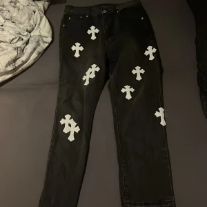 Svarta jeans med vita korsdetaljer - Unika svarta jeans med vita korsapplikationer på både fram- och baksida. Byxorna har klassisk femficksdesign och raka ben. Materialet är jeans och de har en cool streetstil som sticker ut. Perfekt för dig som vill ha något annorlunda i garderoben.