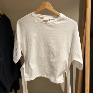 Vit croppad topp med knyt från Nelly - Säljer en vit croppad topp från Nelly i mjuk bomull. Toppen har korta ärmar och snygga knytband i sidorna som kan knytas baktill för en cool detalj. Rund halsringning med ribbad kant. Perfekt för dig som gillar enkel och stilren look.