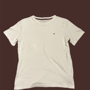 Vit t-shirt från Tommy Hilfiger - Vit t-shirt från Tommy Hilfiger med klassisk rund hals och diskret logga broderad på bröstet. Halsringningen har en snygg randig detalj i rött, vitt och marinblått. T-shirten är gjord i mjuk bomull och har korta ärmar. Perfekt för en clean och stilren look.