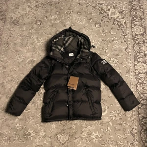 Burberry jacka/väst - Svart pufferjacka från Burberry med huva och klassiskt rutigt foder. Jackan har två framfickor med dragkedja, Burberry London England-patch på ärmen och snygga detaljer. Perfekt för kalla dagar och har en riktigt clean look. Kan även ta av armarna och bli en väst. Helt ny oanvänd